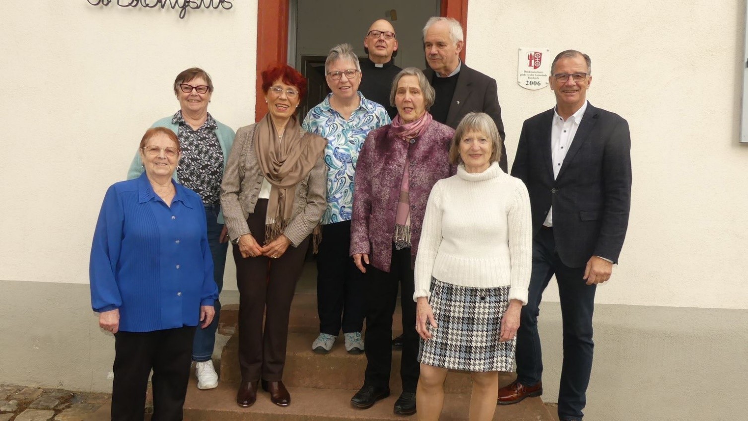 Vor einer hellen Hauswand mit einer geöffneten Tür steht Bürgermeister Winfried Steinmacher, Diakon Hans-Jürgen Siebers und Pfarrer Ralph Senft und sechs Damen.