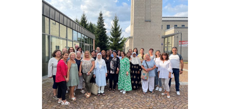 Integrationslotsinnen und -lotsen im Rheingau-Taunus-Kreis vernetzen sich Eine Gruppe von 29 Personen stehen auf Kopfsteinpflaster vor einem aus hellen Steinen gemauertem Gebäude mit Turm. Links ist ein Gebäude mit großer Glasfront und im Hintergrund sind hohe Nadelbäume und ein bewölkter Himmel.