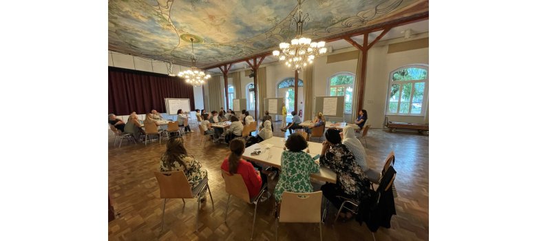 Integrationslotsinnen und -lotsen im Rheingau-Taunus-Kreis vernetzen sich In einem Saal mit zwei Kronleuchtern und blau-weißer Deckenbemalung und Ornamenten sitzen mehrere Personen auf Stühlen an Tischgruppen. Der Saal hat eine Bühne, die mit einem weinroten Vorhang geschlossen ist und an der Wand sind vier große Sprossenfenster und eine große geöffnete Tür. Es stehen vier Leinwände im Saal, auf denen beschriebenes Papier hängt.
