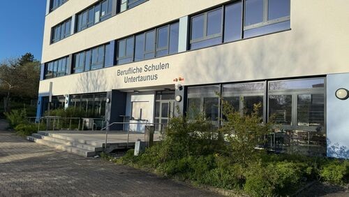 Ein graues Berufsschulgebäude, mit einer breiten Treppe mit drei Stufen am Eingang. Es sind vier Stockwerke mit jeweils einer breiten Fensterfront zu sehen. 