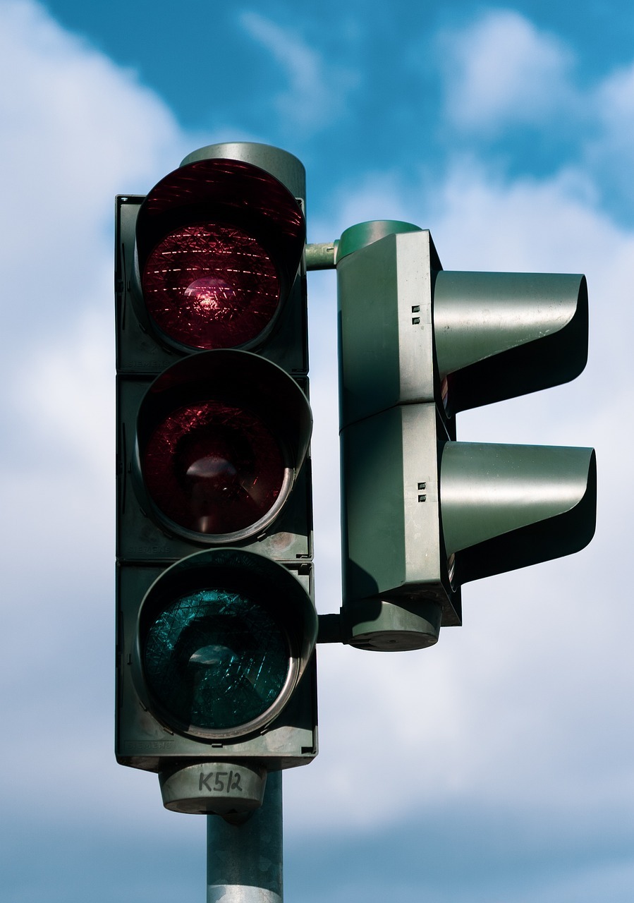 Vor einem blauem Himmel mit weißen Wolken ist eine Ampel, die rot leuchtet. Rechts an der Seite ist die Fußgängerampel mit Blende zu erkennen.