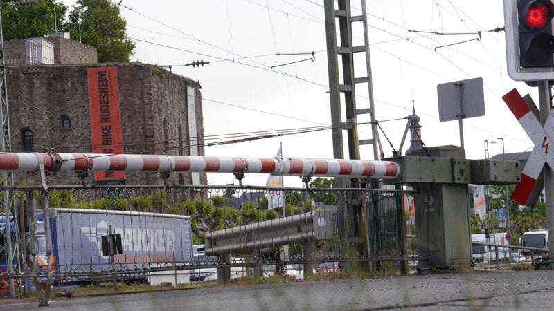 Ein Bahnübergang mit geschlossener Schranke, roter Ampel und Andreaskreuz. Im Hintergrund steht ein blauer LKW und eine Burg mit einem langem roten Banner und ein Kirchturm. Die Stromleitungen der Bahn und ein Mast sind oberhalb der Schranke.