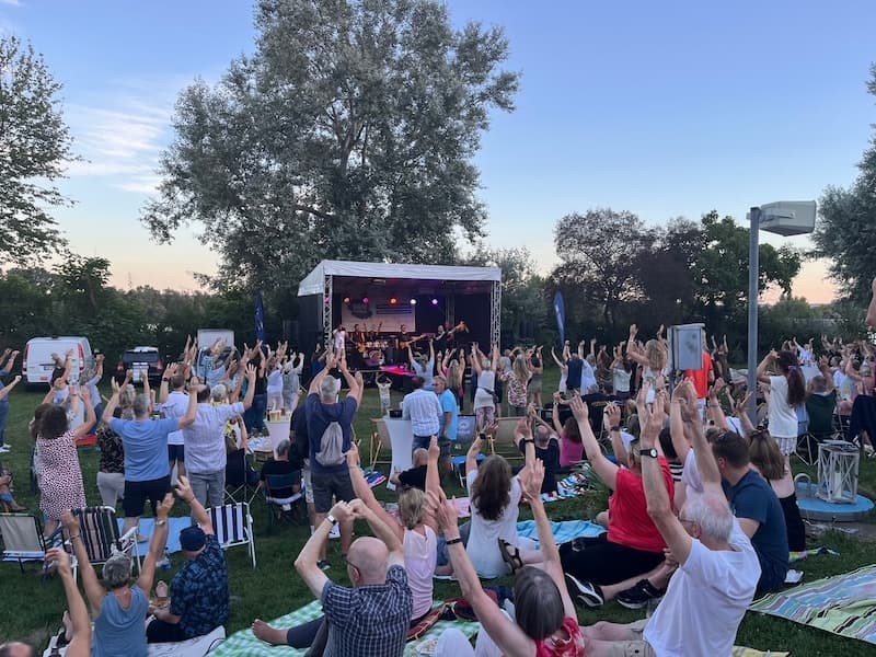 Auf einer Wiese, auf der auch Bäume stehen, ist eine Bühne aufgebaut, auf der eine Band spielt. Auf der Wiese stehen und sitzen Menschen auf Picknickdecken und Campingstühlen. Alle Leute haben die Arme nach oben gestreckt.