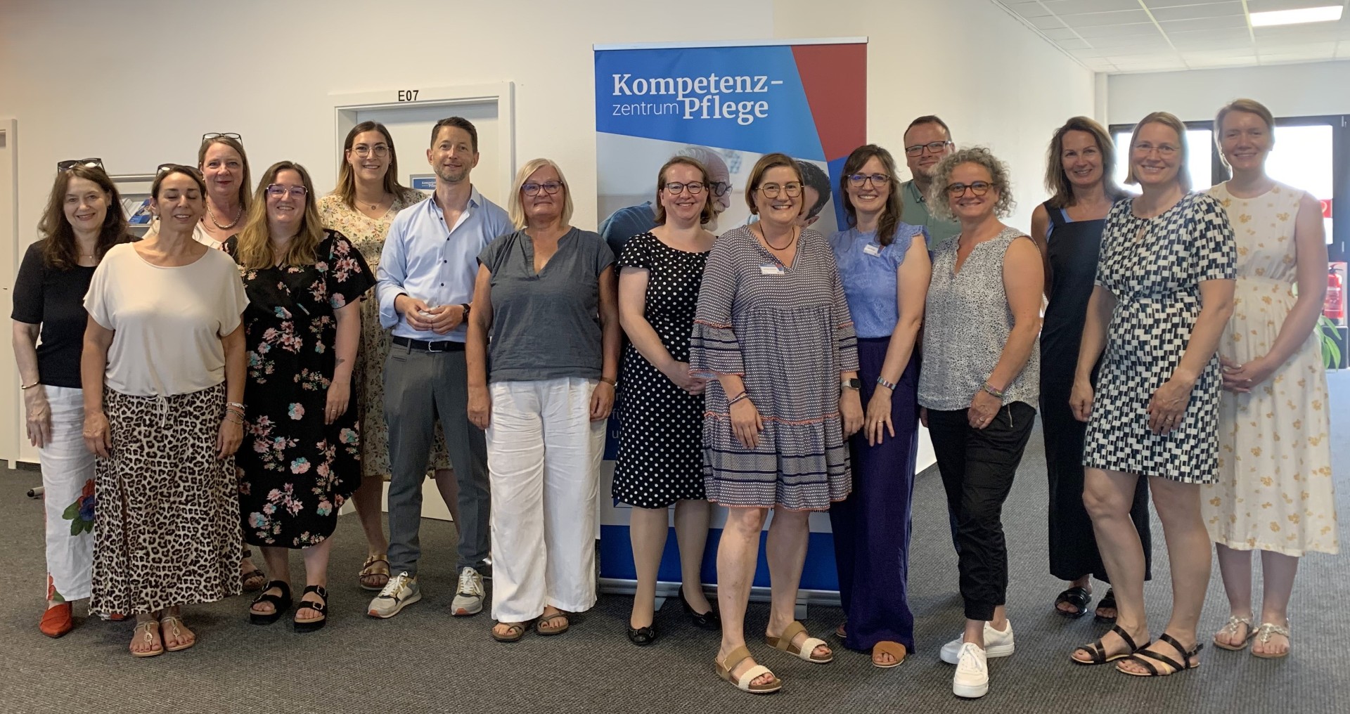 In einem Flur mit Türen und Fenstern stehen 2 Männer und 13 Frauen vor einem Roll-up in sommerlicher Kleidung.