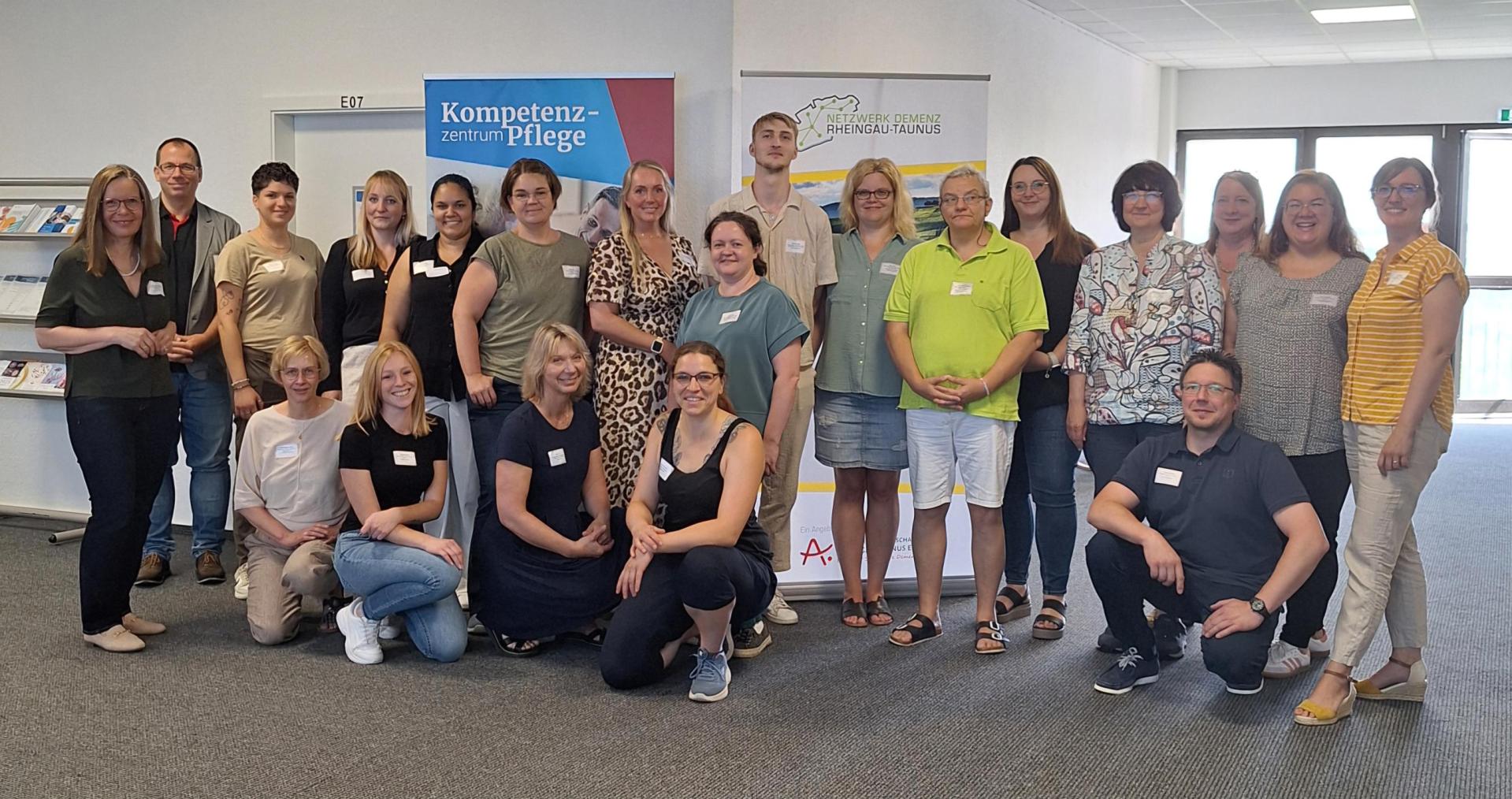 Eine Gruppe, bestehend aus 21 Personen, stehen in einem Flur mit einem Prospektständer und zwei RollUp-Banner. Jeder trägt ein weißes Namensschild.
