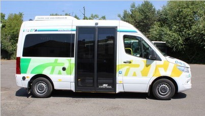 Auf einer Asphaltfläche, vor grünen Laubbäumen und Hecken, steht ein Kleinbus in weiß und großen schwarzen Schiebetüren. Das Logo RTV ist ebenso weiss, aber grün/gelb umrandet.