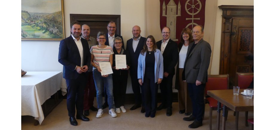 In einem Raum mit zwei alten braunen Türen hängt an der Wand eine Stoffbahn mit einem Wappen in rot, mit beigem Turm und Doppelrad auf beigem Untergrund und ein altes Landschaftsgemälde mit goldenem Rahmen. Davor stehen vier Frauen und 6 Männer überwiegend in dunkeln Anzügen und weißen Hemden.