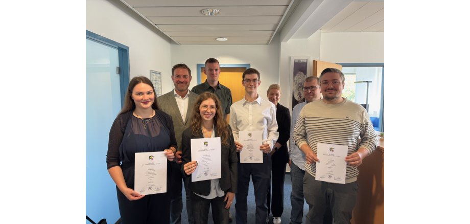 Zwei junge Frauen und zwei junge Männer halten weiße Urkunden in den Händen. Dahinter steht eine Frau und drei Männer. Im Hintergrund links ist eine geschlossene Glastür, geradeaus eine geschlossene Holztür und rechts davon eine geöffnete Holz-Büro-Tür. Die Türrahmen sind blau umrandet und die Wände sind weiß.