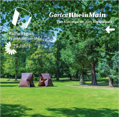 Auf einer Wiese stehen mehrere große, alte grüne Laubbäume und zwei rechteckige braungefleckte Skulpturen, die wie ein aufgestelltes V aussehen.