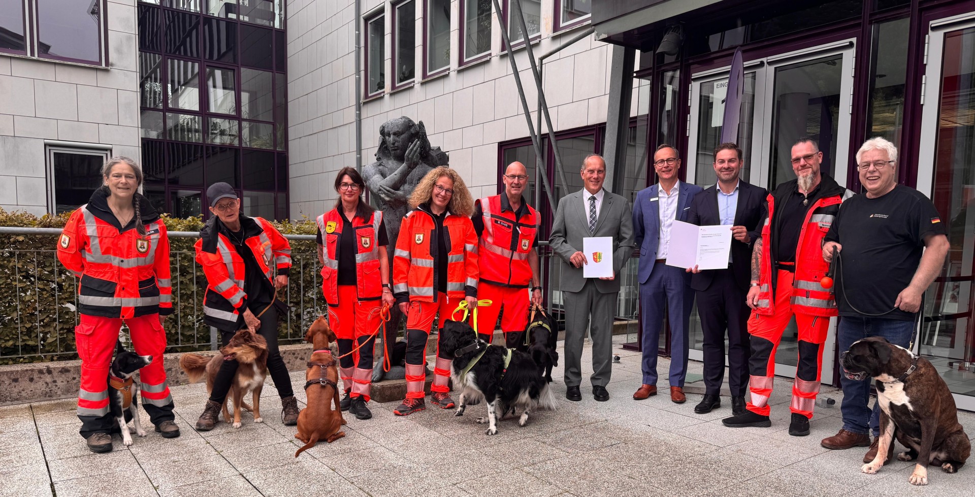 Vor einem Verwaltungsgebäude mit einem großem Glastreppenhaus und vielen Fenstern, stehen 6 Männer und vier Frauen mit sechs Hunden. Drei der Männer haben Anzüge und Hemden an und halten Urkunden in den Händen, der Rest hat orangene Hosen und Jacken mit Reflektierstreifen an. Ganz außen der Mann trägt ein schwarzes T-Shirt und eine blaue Jeans. Im Hintergrund ist eine Statue aus Stein zu erkennen. 