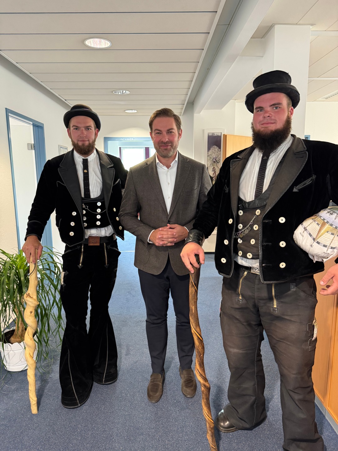 Landrad Zehner steht im Anzug zwischen zwei Gesellen auf der Walz. Sie tragen den schwarzen Hut, die Traditionskleidung in schwarz mit weißen Knöpfen, die Weste und das weiße Hemd. Beide tragen einen langen Bart und haben geschwungene Wanderstöcke inn der Hand.