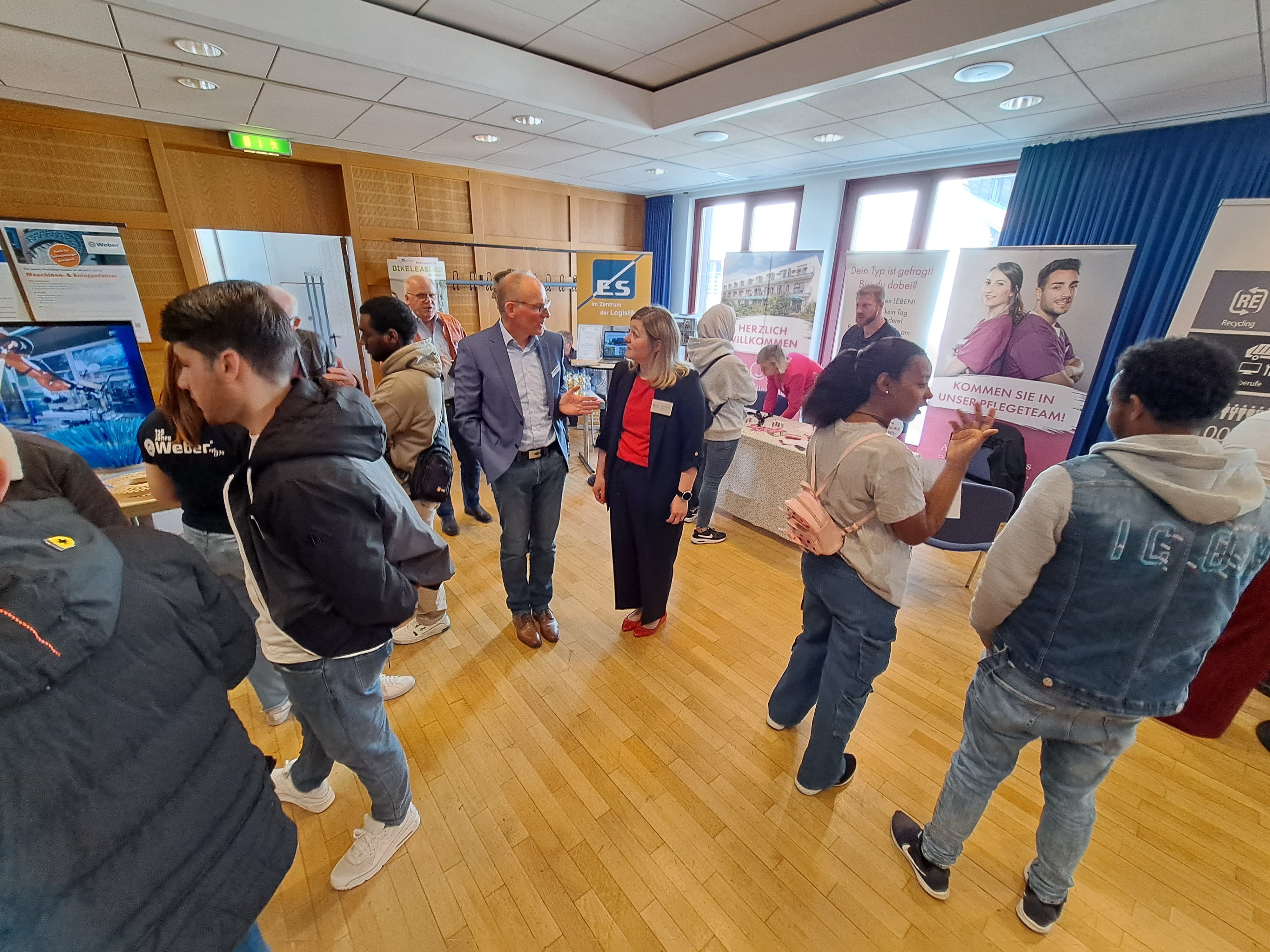 In einem Raum stehen mehrere Menschen und sprechen miteinander. Es sind InfoTische mit Flyern aufgestellt, hinter denen Roll-Ups mit Werbung für Firmen für einen Ausbildungs- oder Arbeitsplatz stehen.