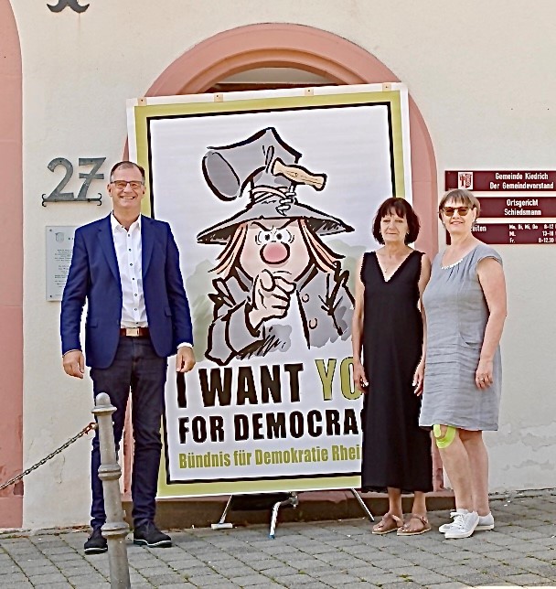 Bürgermeister Steinmacher und zwei Frauen in Kleidern stehen vor dem Rathaus und vor einem Plakat, auf dem ein gezeichneter Mann mit grauem Schlappgut und einem Korkenzieher an der Hutkrempe und einer grauen Jacke abgemildet ist. Er zeigt mit dem Finger auf den Betrachter, schaut grimmig und unter ihm steht "I WANT YOU FOR DEMOCRATY".  