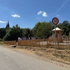 Blick auf eine Straße, mit einem 10 km/h Verkehrsschild, einen Holzzaun und dahinter einem Spielplatz mit mehreren Menschen und einem Holzturm mit Rutsche. Dahinter sind Bäume und der Kirchturm der Kiedricher Kirche zu erkennen. Der Himmel ist bewölkt und hat wenige weiße Wolken.  