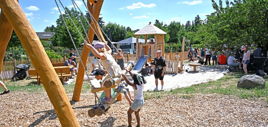 Auf einer Mehrgenerationschaukel sitzen mehrere Kinder und ein Kind schubst an. Dahinter ist eine Banksitzgruppe und eine Rutsche und ein Holzturm zu sehen. Es ist ein großer Sandkasten um die Rutsche und Holzspäne um die Schaukel. Auf dem Spielplatz sind viele Menschen und im Hintergrund viele Bäume.