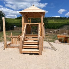 In einem großem Sandkasten steht ein Holzturm mit einer Treppe und einem Kletternetz sowie einem Zaun und einem Steuerrad. Im Hintergrund ist eine Sitzgarnitur und eine Wippe zu erkennen. Außerhalb des Zaunes sind Weinberge, oberhalb der Weinberge Wald und ein leicht bewölkter Himmel.