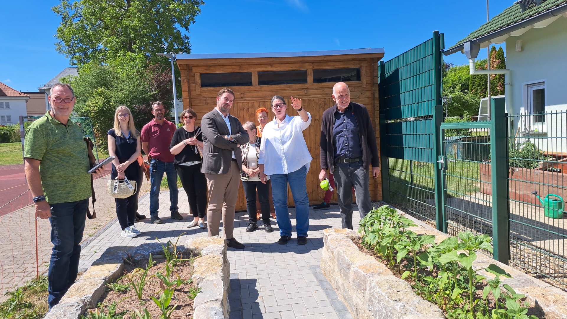 Hinter zwei langen Hochbeten mit Steinumrandung und Bepfalnzung, stehen vier Männer und fünf Frauen vor einer Holzhütte. In der näheren Umgebung sind weitere Häuser erkennbar und grüne Bäume sowie ein blauer Himmel ohne Wolken.