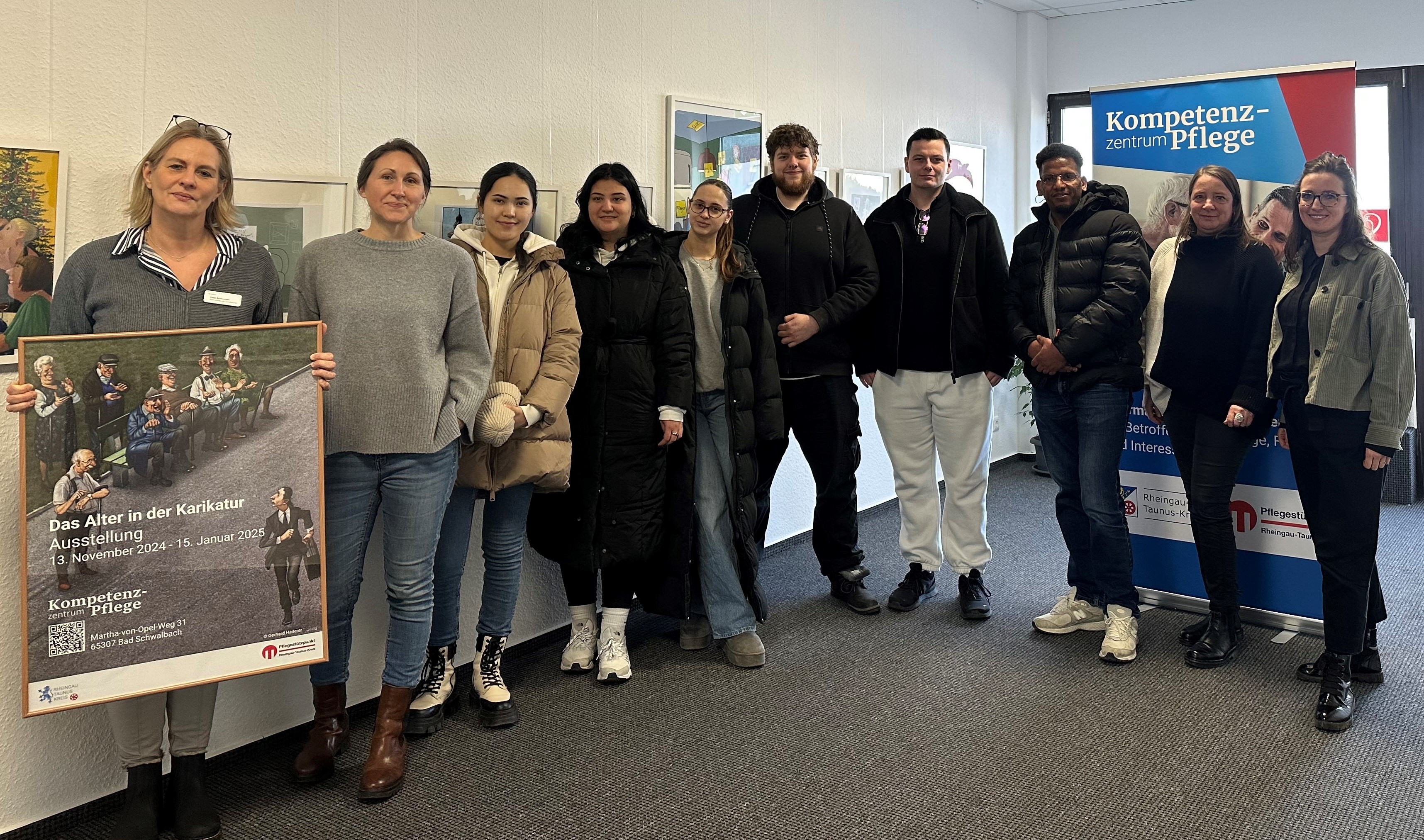 Sieben Frauen und drei Männer stehen vor einem Banner des Kompetenzzentrum Pflege. Die Frau ganz links hält einen Rahmen mit einem Werbeplakat für die Ausstellung "Das Alter in der Karikatur" in der Hand. 