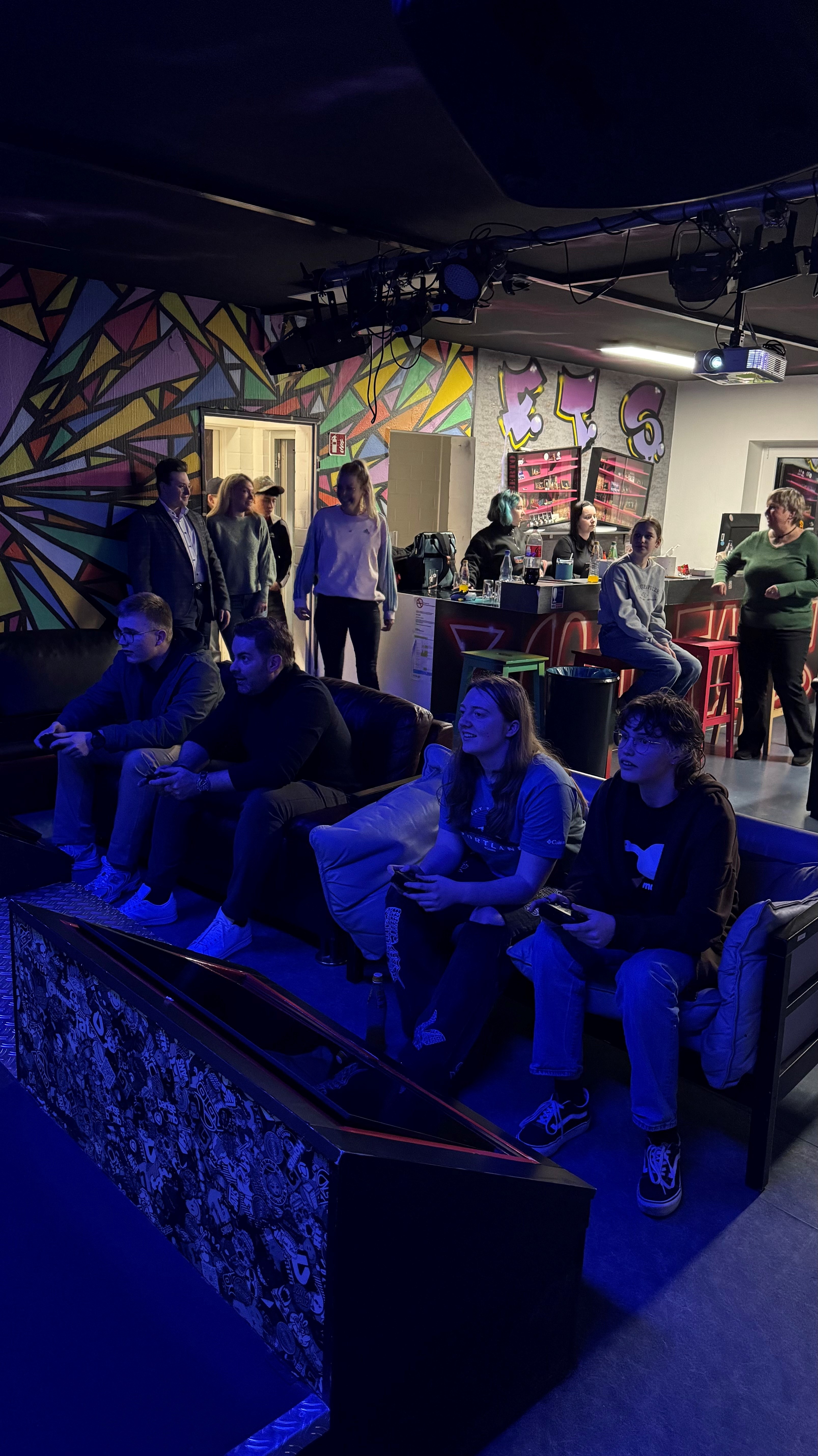 In einem Raum sind Grafitti, lauter bunte Dreiecke mit schwarzen Rahmen und Buchstaben an der Wand. Im Hintergrund ist eine Bar und im Vordergrund zwei Sofas, die mit vier Personen besetzt sind und Gamecontroler in den Händen halten. Es schimmert bläuliches Licht und es sind mehrere Personen im Raum.