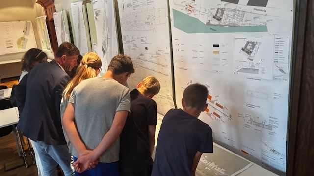 Vor mehreren Pinwänden, auf denene diverse beschriftete Blätter gepinnt wurden, stehen mehrere Kinder und Erwachsene, in Summe 7, die sich vorbeugen um sich etwas anzuschauen.