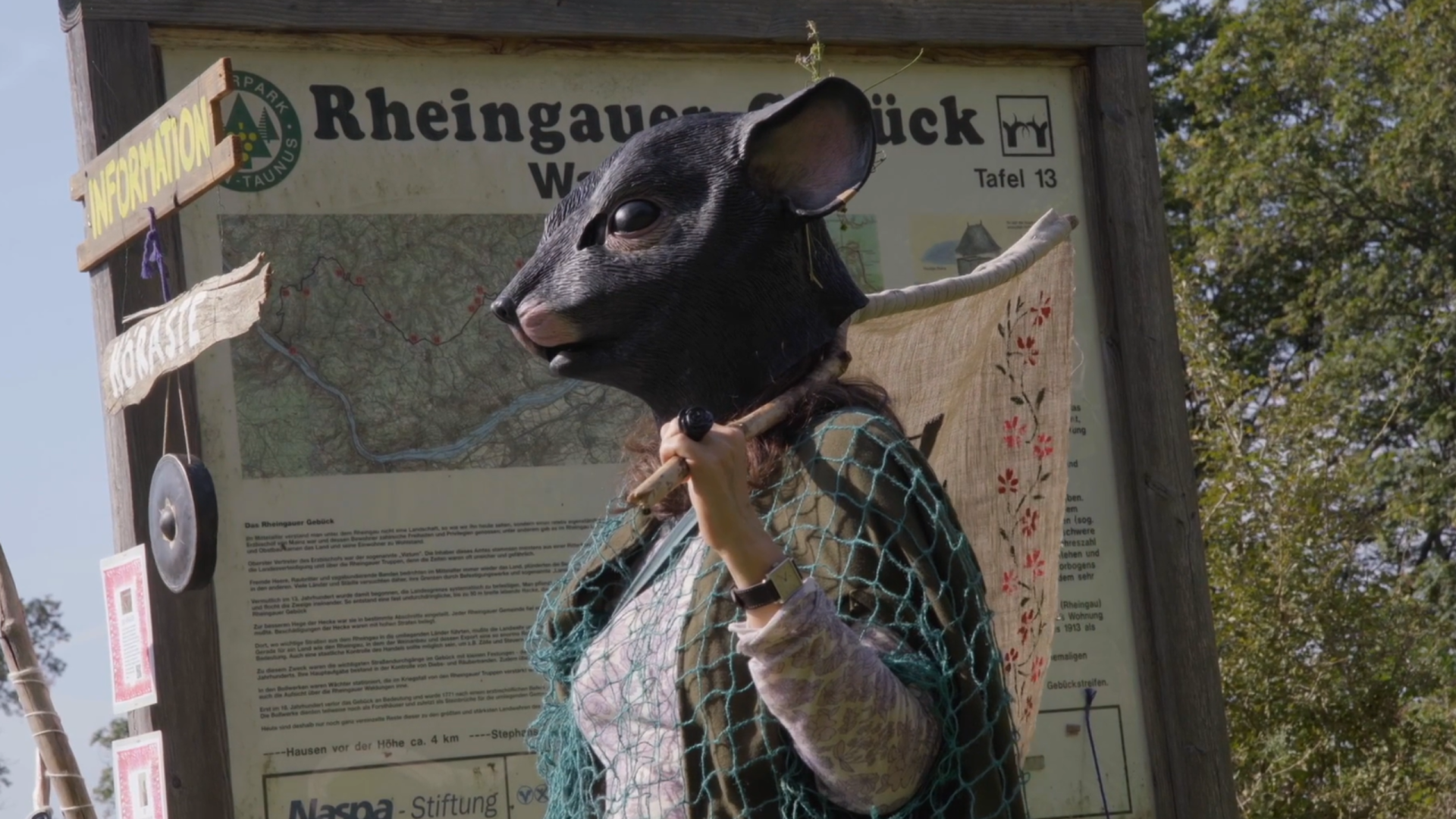 Ein Mensch hat einen lila-weißen Pullover an und darüber eine grüne Strickweste. Darüber ist ein blaues Fischernetz gezogen. In der Hand wird ein Stock gehalten, der auf der Schulter liegt und an dem eine beige Fahne hängt. Am Rand der Fahne sind rote- und rosa-farbene Blüten mit grünen Blättern gedruckt. Auf dem Kopf wird eine Maske aus Kunststoff getragen, die wie eine Maus aussieht. Sie bedeckt den kompletten Kompf. Im Hintergrund ist eine Informationstafel zum Rheingauer Gebück und daneben sind Laubbäume.