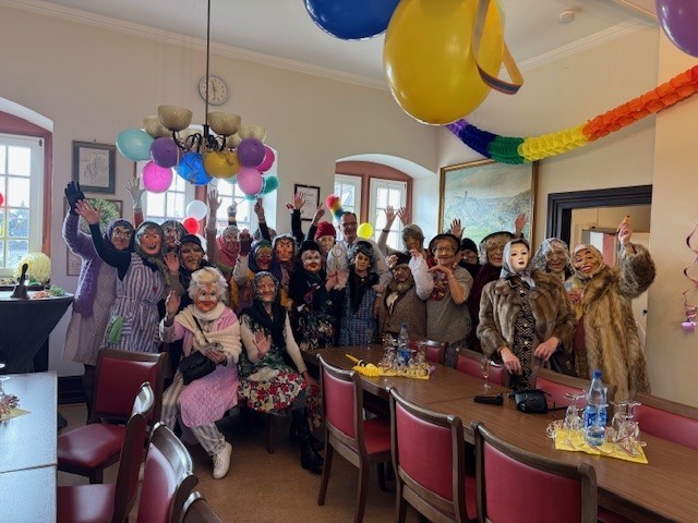 Im dem mit Luftbarlons und Girlanden geschmückten Rathaussaal,  steht der Bürgermeister, umringt von 23 Frauen, die als alte Damen verkleidet sind. Sie haben Masken auf und Mäntel oder Kittelschürzen an.