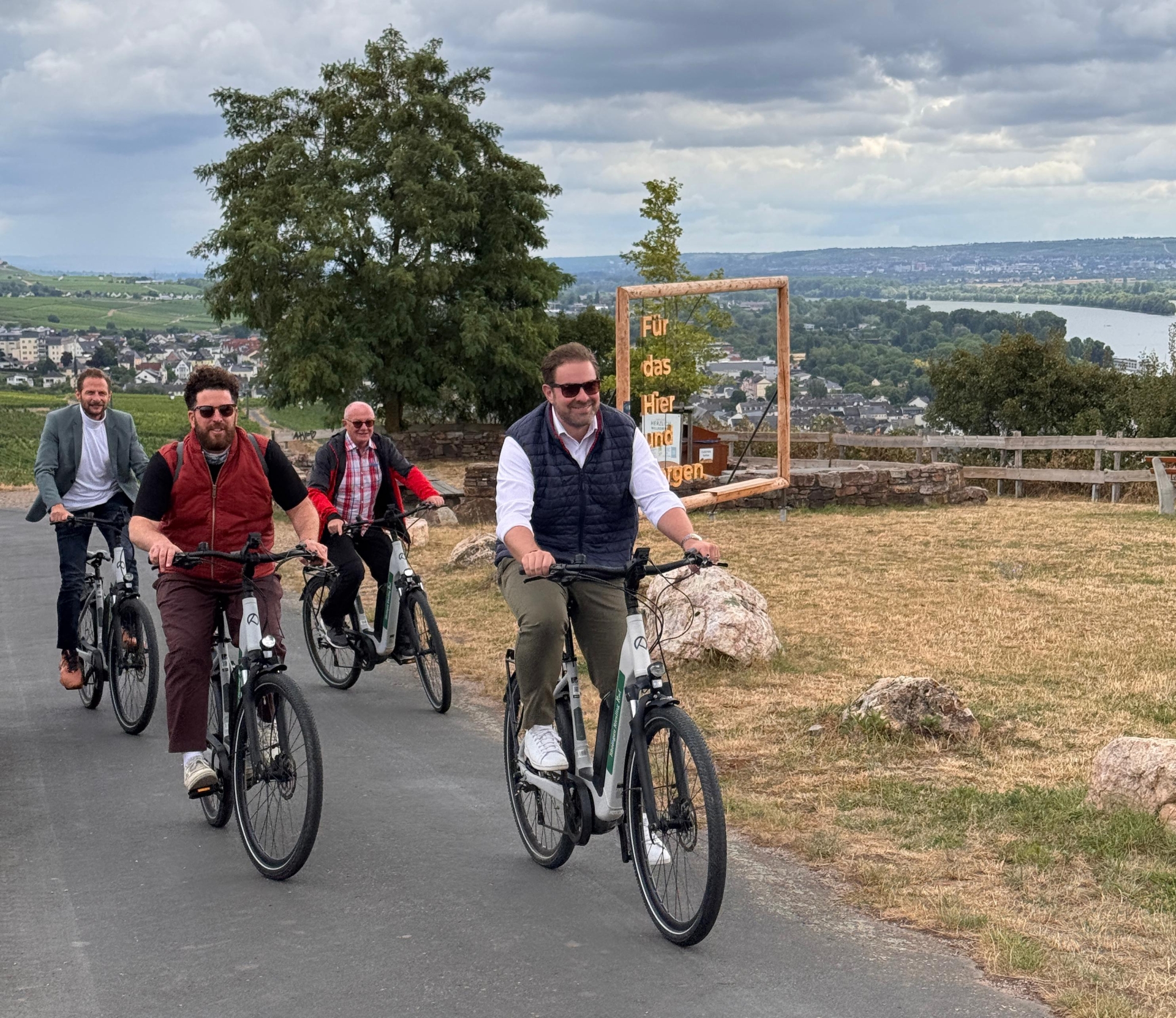 Vier Herren sitzen auf Fahrrädern und fahren eine asphaltierte Straße hoch. Am Straßenrand ist eine Wiese, die gelblich ist und große Steine liegen mit Abstand in einer Reihe. Im Hintergrund ein Fotorahmen aus Holz und ein großer Laubbaum und der Blick auf das Rheintal.