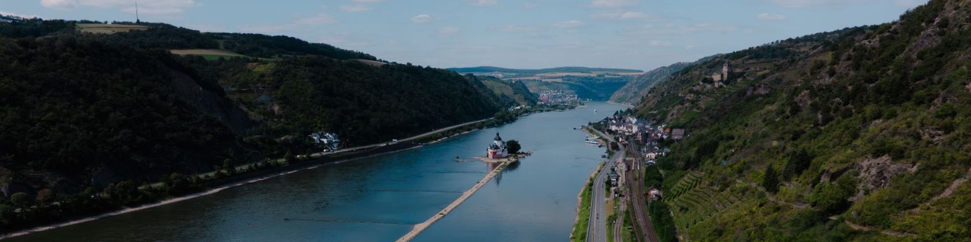 Blick auf das Rheintal und dem Rhein mit den Hängen links und rechts mit Wäldern, Weinbergen, Feldern, Burgen und im Rhein eine weitere Burg und einer langen Sandbank und Anlegern und eine Fähre. Der Himmel ist etwas bewölkt.