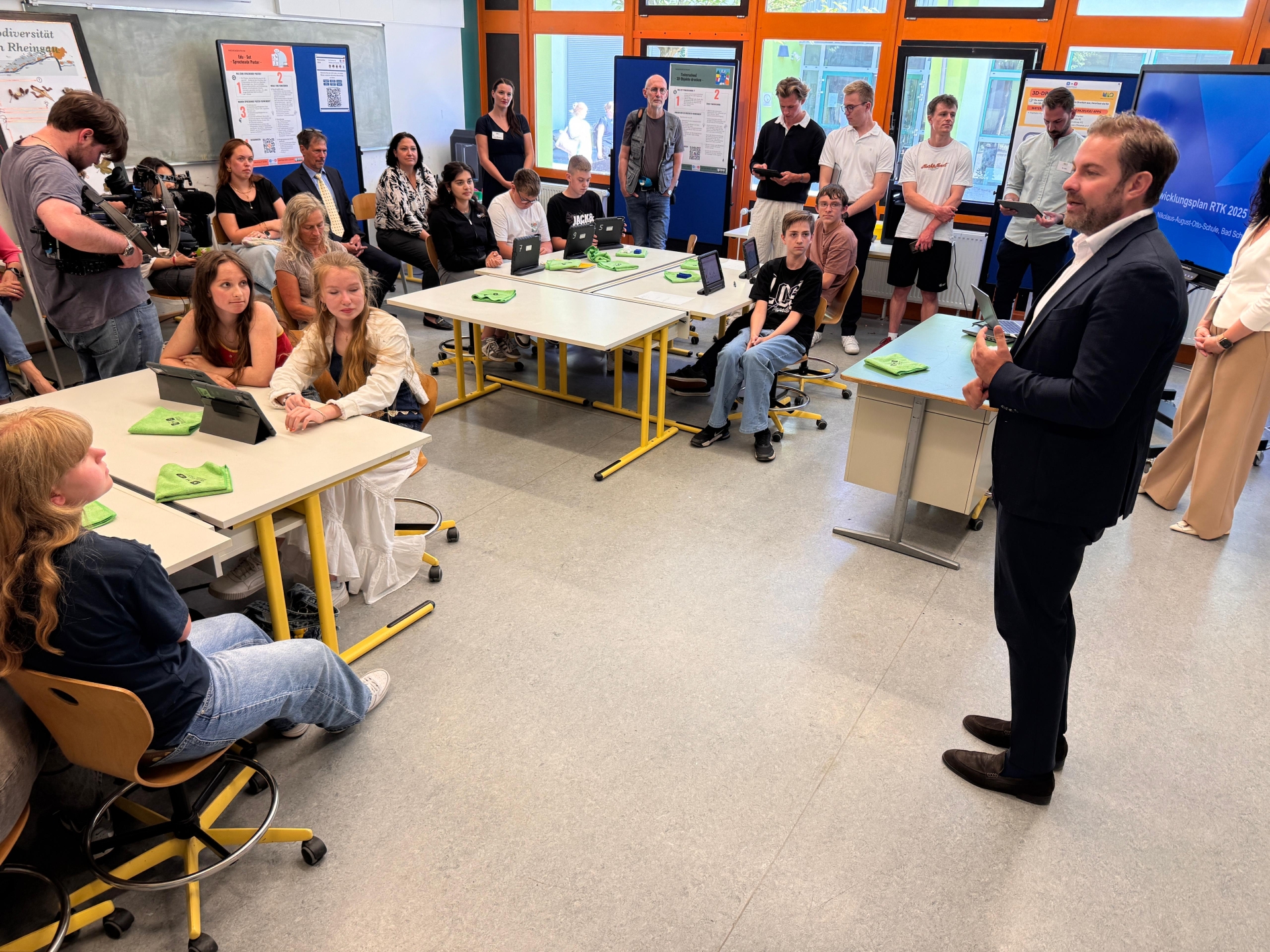 In einem Klassenraum sitzen mehrere Schülerinnen und Schüler an Tischen. Vor ihnen steht jeweils ein Tablett. Außen rum sitzen und stehen weitere Personen. Vor den Wänden und der Fenstefront sind Stellwände mit Plakaten und eine Bildschirm. Vorn steht Landrat Zehner im Anzug und weißem Hemd.