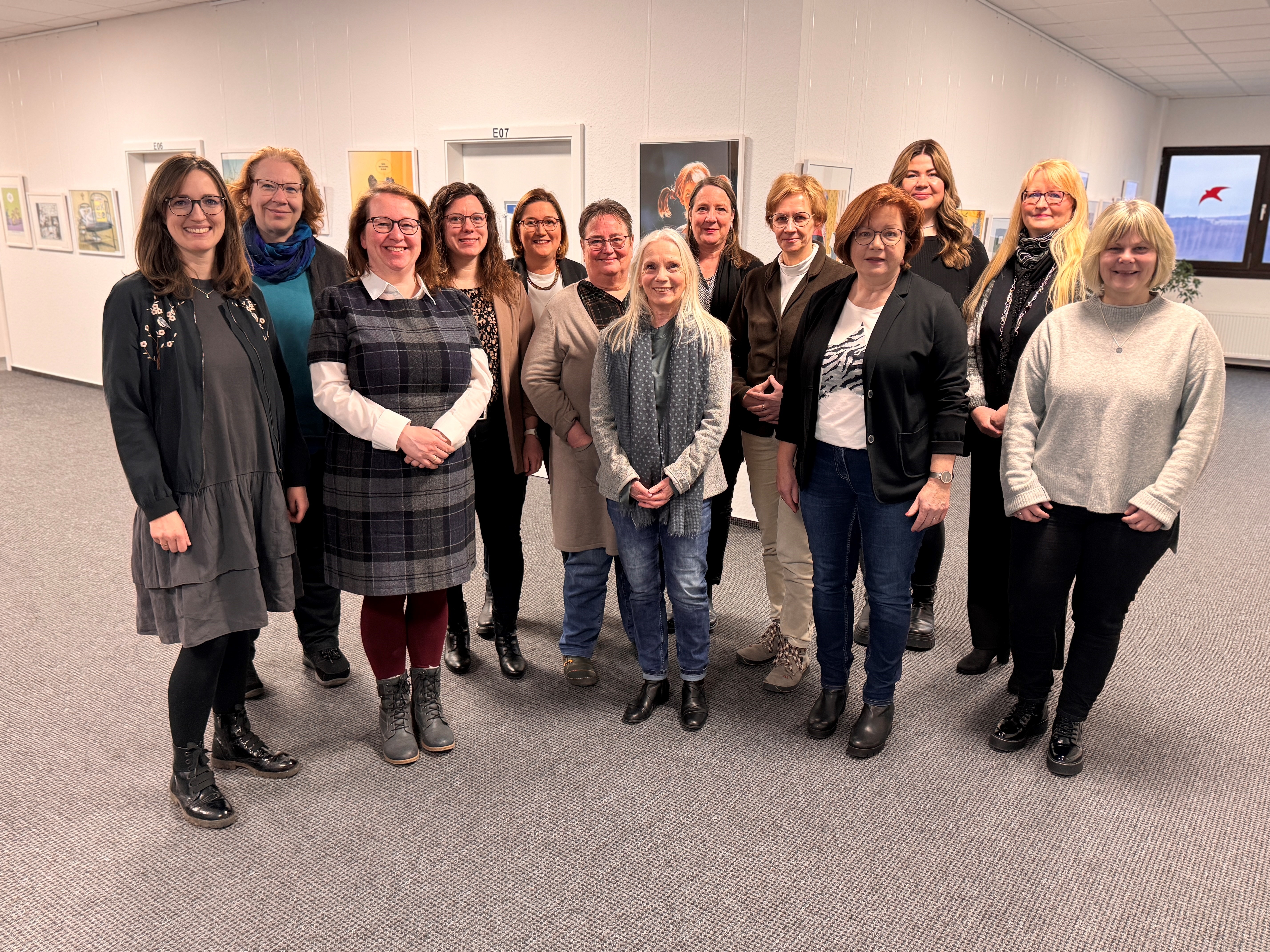 In einem L-förmigem Flur mit Bildern an der Wand und zwei Türen an der einen Seite und einem Fenster mit einer Heizung darunter stehen nebeneinander 13 Frauen.