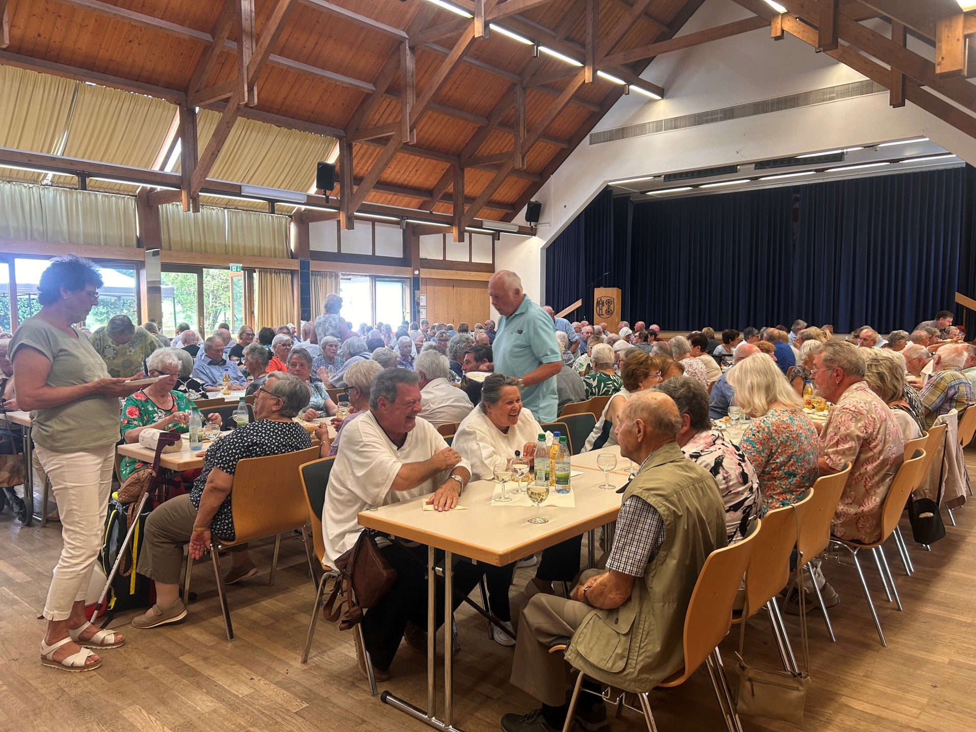 In einem großem Saal, in dem sich eine Bühne mit einem blauem, dicken Bühnenvorhang befindet, stehen Tischen und Stühle. Auf den Stühlen sitzen viele Männer und Frauen.