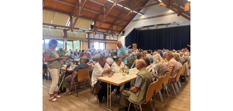 In einem großem Saal, in dem sich eine Bühne mit einem blauem, dicken Bühnenvorhang befindet, stehen Tischen und Stühle. Auf den Stühlen sitzen viele Männer und Frauen.