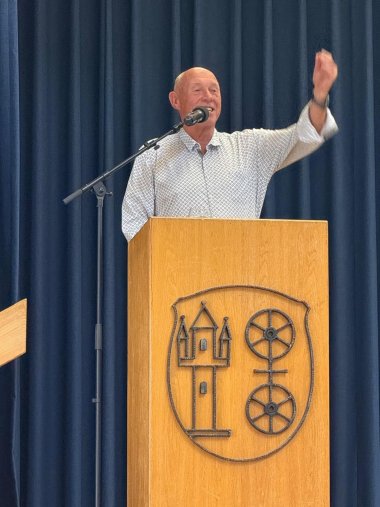 Hinter einem braunem Pult, auf dem mit Metall das Kiedricher Wappen mit Doppelrad und dem Burgturm abgebildet ist, steht ein Mann mit weißem Hemd, weißen Haaren, der seinen linken Arm mit geballter Faust motiviert nach oben streckt. Im Hintergrund ist ein blauer, dicker Bühnenvorhang.