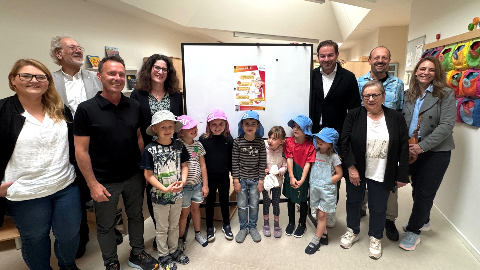 In einem Flur eines Kindergartens stehen 4 Frauen und 4 Männer und 7 Kinder, vor einem Board, an dem ein Plakat hängt. Die Kinder haben beige, rosa und blaue Sonnenhüte auf.