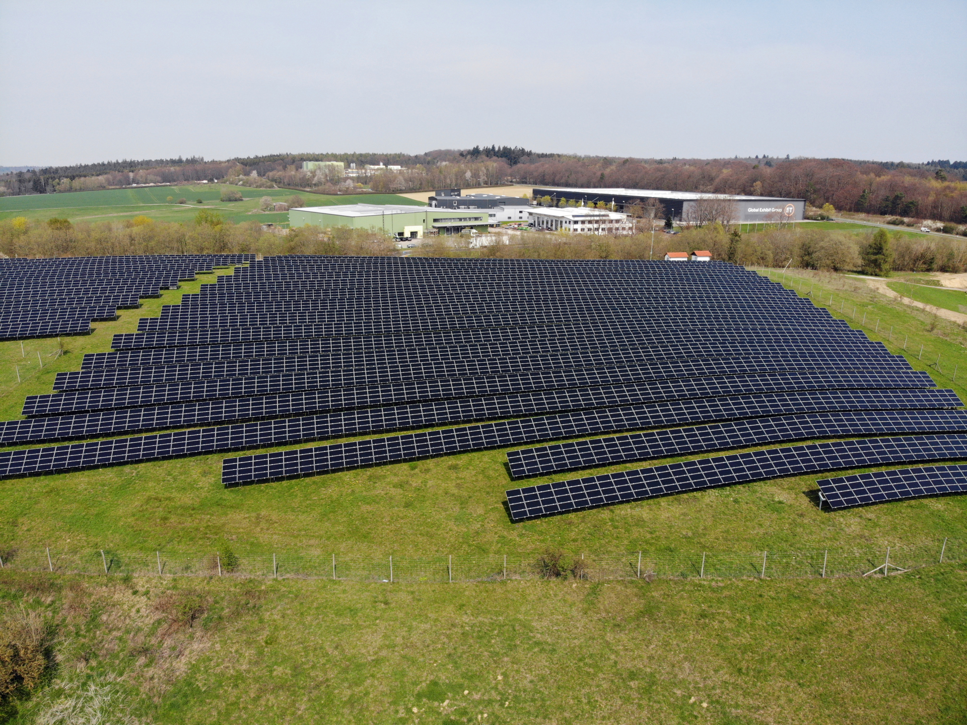 Aus der Luftperspektive wird ein Solarpark, der aus mindestens 19 Reihen und einer zweiten Fläche mit mindestens 11 Reihen Panelen besteht, dargestellt. Rund herum ist Wiese und im Hintergrund mehrere große Firmenhallen. Im Horizont ist Wald zu sehen. Der Himmel ist grau.