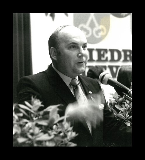 Ein Mann mit kurzen Haaren im dunkelm Anzug und Kravatte und weißem Hemd steht hinter einem Mikrofon. Links und rechts unten sind Blätter und im HIntergrund ein Vorhang und eine große Leinwand mit einem Wappen und Buchstaben. Das Bild ist in schwarz-weiß und hat einen schwarznen, breiten Rahmen.  