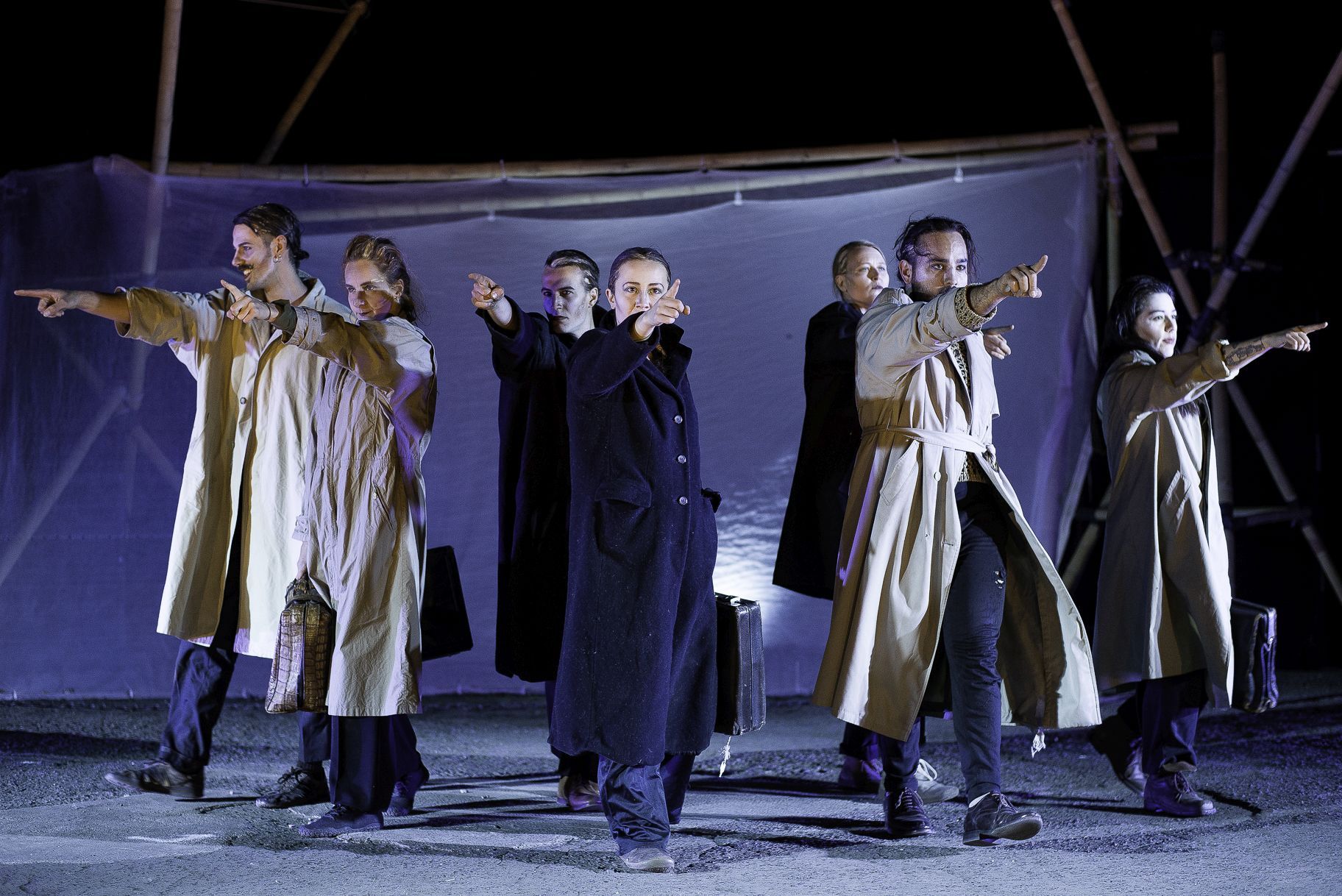 Auf einem grauen Boden stehen drei Männer und vier Frauen. Alle tragen helle oder dunkle, lange, geschlossene Mäntel und haben eine Aktenkoffer in der einen Hand. Die andere Hand ist ausgestreckt und mit dem Zeigefinger wird auf etwas gezeigt. Im Hintergrund ein dunkelblauer, durchsichtiger Banner, der an Stangen befestigt ist.