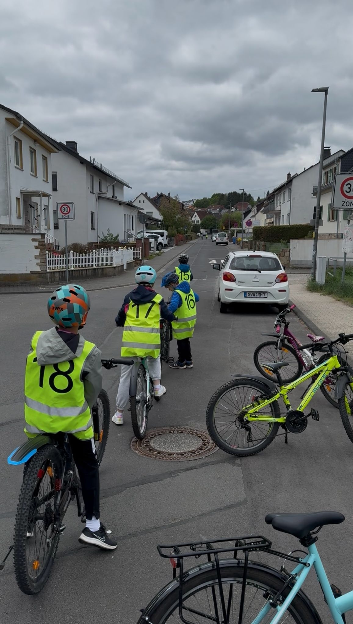 Auf einer Straße mit einer 30er-Zone im Wohngebiet parkt ein weißes Auto. Auf der Straße sieht man vier Kinder. Drei Kinder sitzen auf Ihren Fahrrädern und ein Kind steht neben seinem Fahrrad. Sie tagen Helme auf dem Kopf und gelber Warnfeste mit Zahlen auf dem Rücken. Am Straßenrand stehen drei abgestellte Fahrräder.