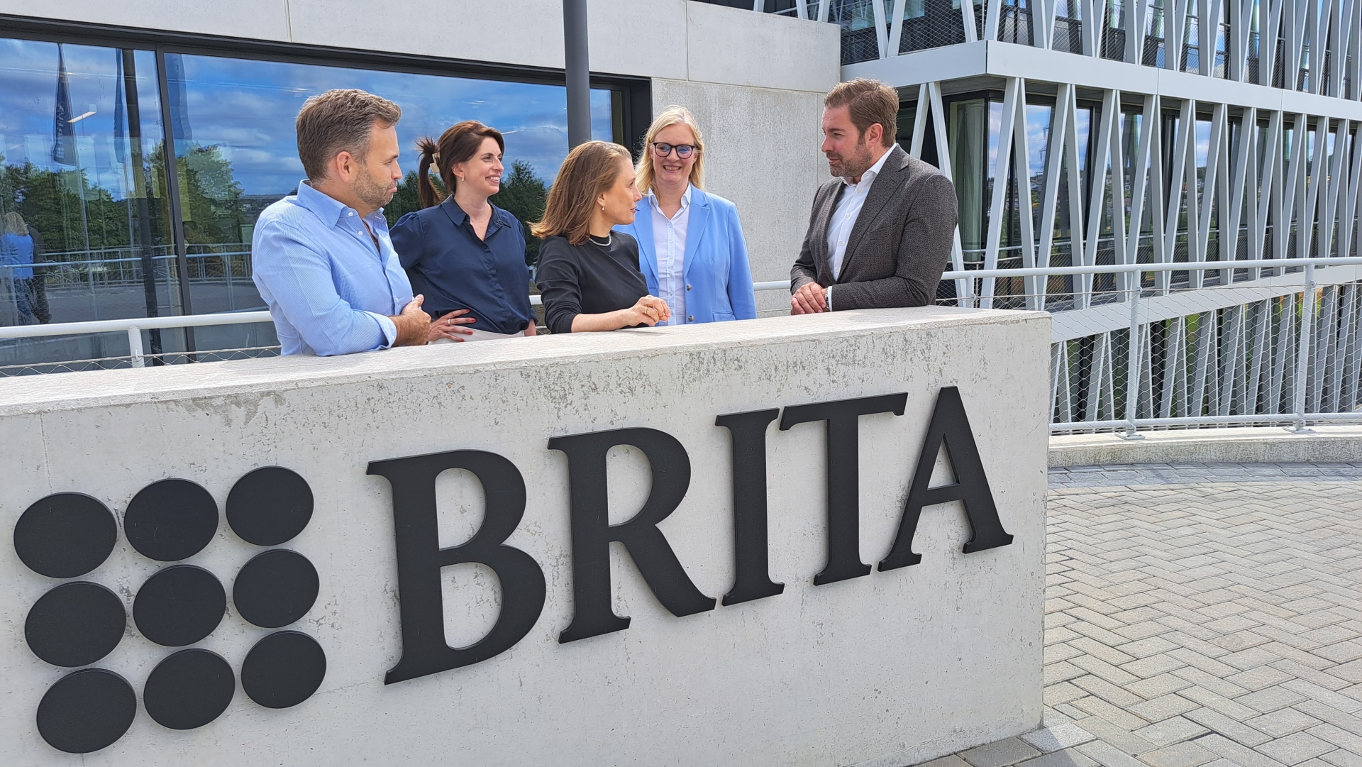 Hinter einer Betonwand, auf der das Firmenlogo von Brita geschrieben ist, stehen zwei Männer und drei Frauen. Dahinter ist eine breite Glasfront von Betonsteinen eingefasst und rechts ist ein großes Gebäude mit Stahlträgern als Dreiecken vor den Fenstern.