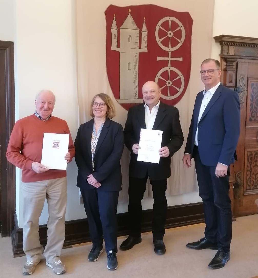 Vor der Fahne mit dem Kiedricher Wappen, dem Scharfenstein u nd dem Doppelrad in beige, auf rotem Untergrund, stehen v.l.n.r. Wilfried Ringel im orangenem Pullover und beiger Hose und Kerstin Merno, Georg Sohlbach, Bürgermeister Winfried Steinmacher, alle im dunklem Anzug mit weißem Hemd/Bluse. Herr Ringel und Herr Sohlbach haben Urkunden in der Hand.