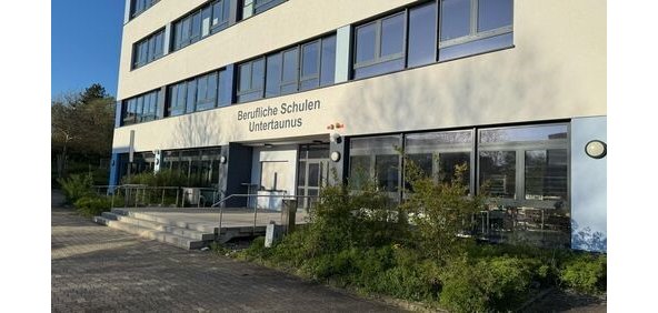 Ein graues Berufsschulgebäude, mit einer breiten Treppe mit drei Stufen am Eingang. Es sind vier Stockwerke mit jeweils einer breiten Fensterfront zu sehen. 