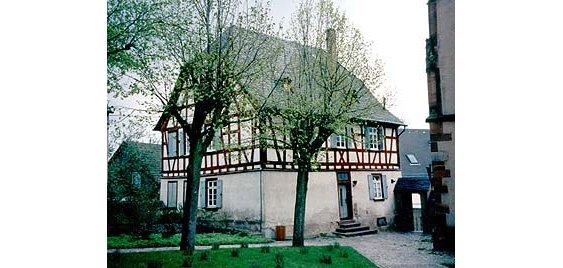 Ein Fachwerkhaus, weiß mit braunen Balken. Im Vordergrund drei Bäume, bei denen die Äste abgesägt wurden, eine Wiese mit Grabsteinen, stehende Kreuze und ein geplasterter Weg. Rechts ein Teil der St. Michaelskapelle in mit großen Fenstern und Verzierungen aus Sandstein. 