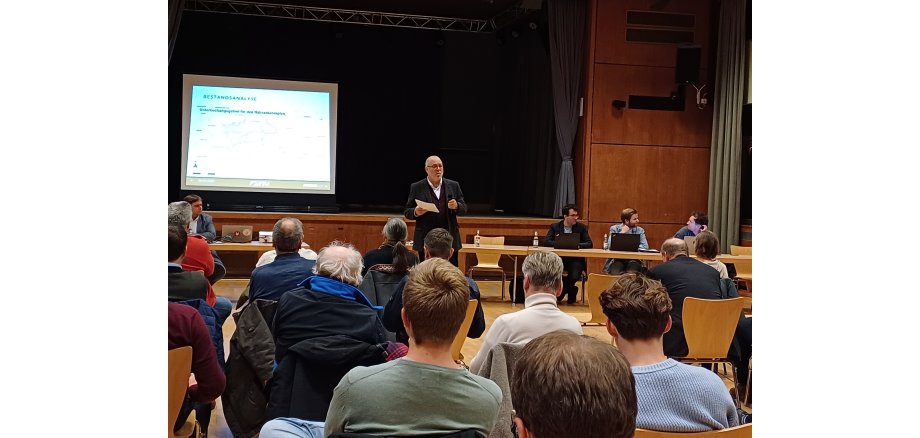 Auf einer Bühne mit einem dunklem Hintergrund  steht eine Leinwand, auf die eine Präsentation gezeigt wird. Davor stehen Tische, an denen vier Personen mit aufgeklappten Laptops ins Publikum schauen. Das Publikum sitzt gegenüber. Ein Redner im dunklem Anzug und weißem Hemd steht mit einem Zettel in der Hand vor dem Publikum. 