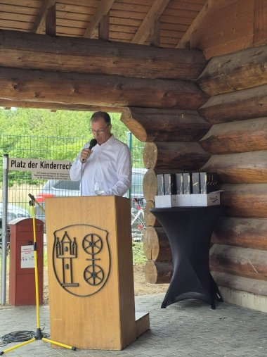 Jubiläum 30 Jahre Kita Hickelhäusje An einem Rednerpult aus Holz mit dem Gemeindewappen steht Bürgermeister Steinmacher mit einem weißen Hemd und Mikrofon in der Hand. Dahinter ein Bistrotisch mit Weinpräsenten und ein großer roter Briefkasten. Im Hintergrund sind grüne Blätter und das Holz eines Baumstammhauses zu erkennen.