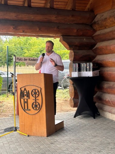 An einem Rednerpult aus Holz mit dem Gemeindewappen steht ein Mann mit einem weißen Hemd und Mikrofon in der Hand. Dahinter ein Bistrotisch mit Weinpräsenten und einem Straßenschild. Im Hintergrund sind grüne Blätter und das Holz eines Baumstammhauses zu erkennen.