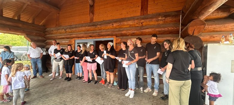 Vor einem Blockhaus aus Holz stehen Erzieher, Erzieherinnen mit schwarzen Shirts und weißen Zetteln in der Hand vor einem Blockhaus aus Holz. Ebenso sind Kinder dabei.