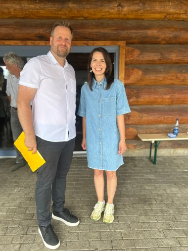Ein Mann in grauer Hose und weißem HEmd und gelber Mappe in der Hand steht neben einer Frau mit braunen schulterlangen Harren und Jeanskleid vor einem Baumstammhaus.