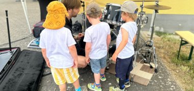 Vor einem Schlagzeug stehen drei Jungs im weißem T-Shirt und kurzen hosen und ein Mann im schwarzem Shirt kniet dazwiwschen. Im HIntergrund steht ein Stehtisch und eine Bank vor einer gelben Wand.