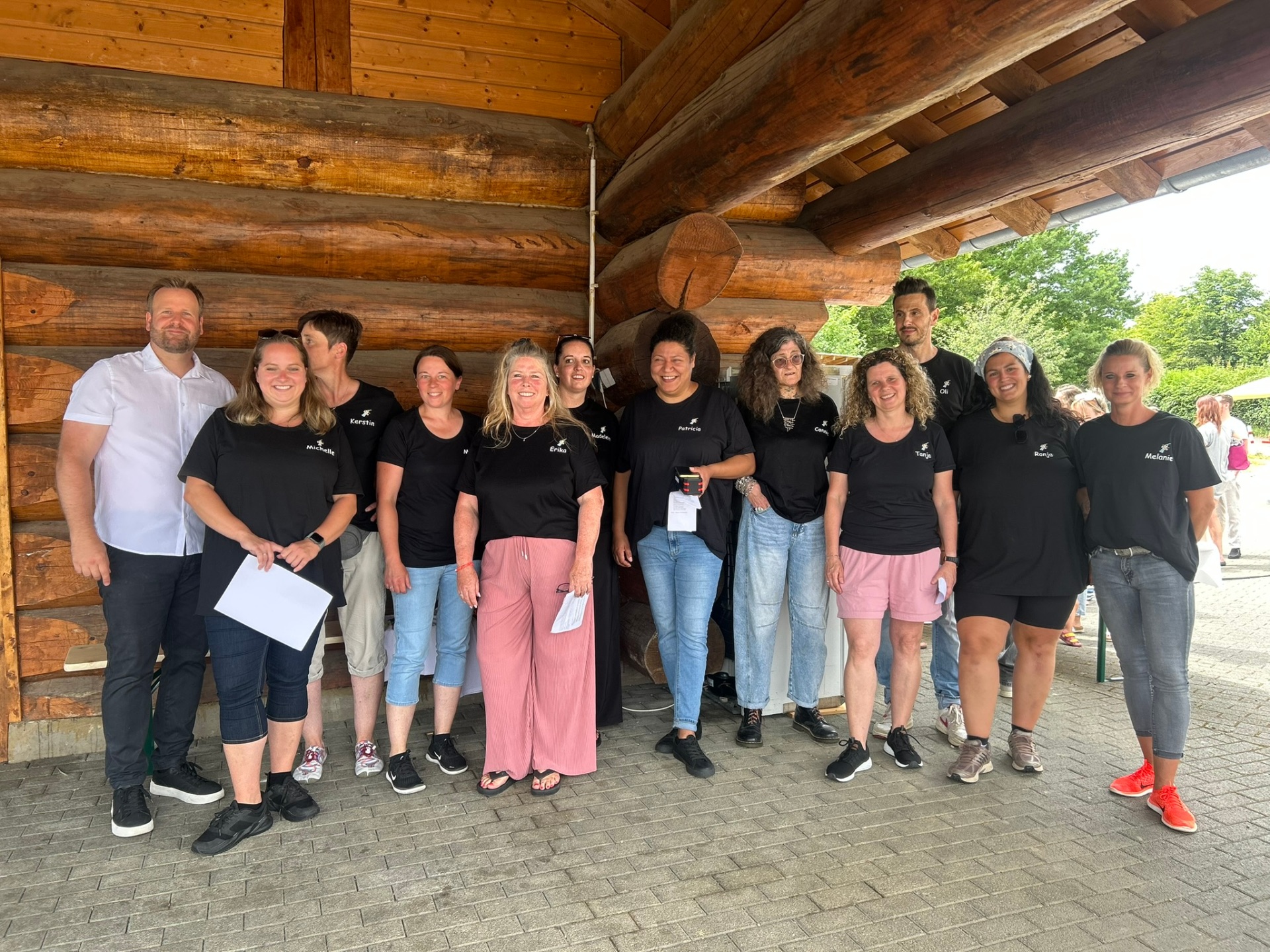 Vor einem Blockhaus aus Holz stehen zwei Erzieher und 10 Erzieherinnen mit schwarzem Shirt und einem weißem Hemd. 