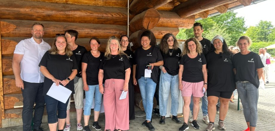 Vor einem Blockhaus aus Holz stehen zwei Erzieher und 10 Erzieherinnen mit schwarzem Shirt und einem weißem Hemd.